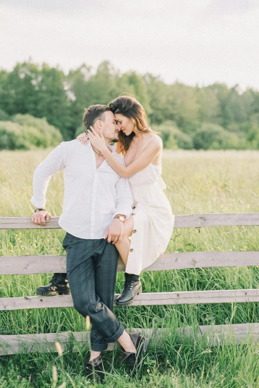 a man and woman sitting on top of a wooden fence, in a grass field, white hue, 2019 trending photo, romantic lead