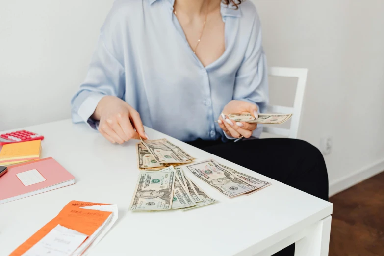 a woman sitting at a table with a stack of money, pexels contest winner, 🦩🪐🐞👩🏻🦳, wearing a light blue shirt, avatar image, candid photo