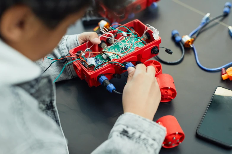 a person working on an electronic device on a table, by Jakob Gauermann, only one robot kid on the ground, red and black robotic parts, 🦩🪐🐞👩🏻🦳, prototype car