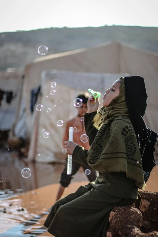 a woman blowing bubbles while sitting on a rock, by Youssef Howayek, trending on unsplash, conceptual art, in a war - torn desert village, a person at a music festival, square, 14 yo berber boy