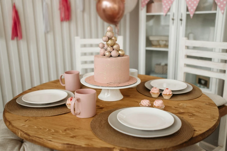 a pink cake sitting on top of a wooden table, by Alice Mason, pexels contest winner, cups and balls, dining table, white clay, round base