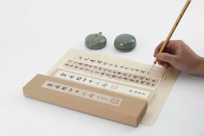 a person holding a pencil and writing on a piece of paper, an album cover, inspired by Tang Yin, mingei, celadon glaze, box, with some hand written letters, horizontal orientation
