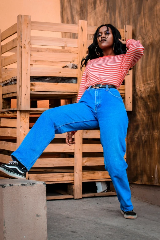 a woman leaning on a stack of wooden pallets, by Everett Warner, trending on pexels, blue long pants and red shoes, woman in streetwear, ( ( dark skin ) ), cropped wide sleeve