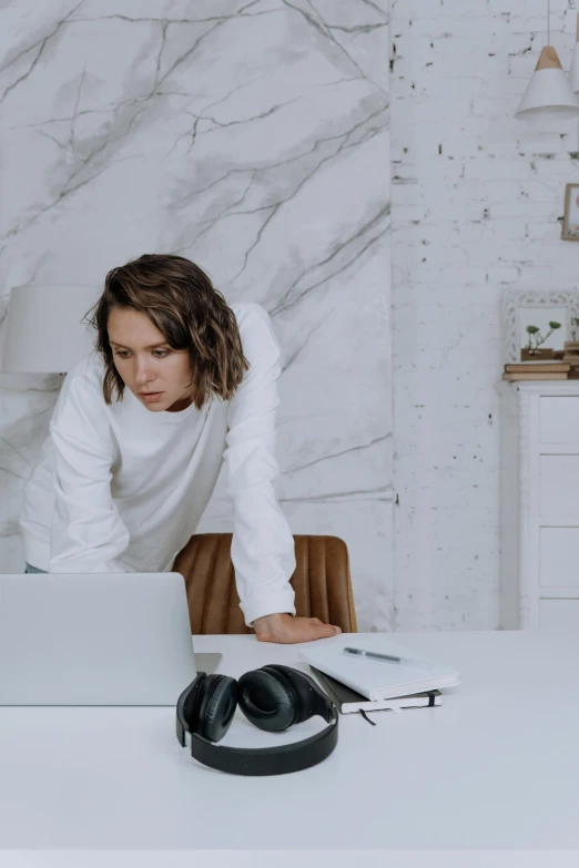 a woman sitting at a table with a laptop and headphones, trending on pexels, arbeitsrat für kunst, white marble interior photograph, wearing a white sweater, serious business, bending down slightly