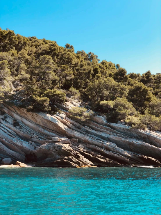 a person riding a surf board on top of a body of water, calanque, trees and cliffs, deep colours, panoramic