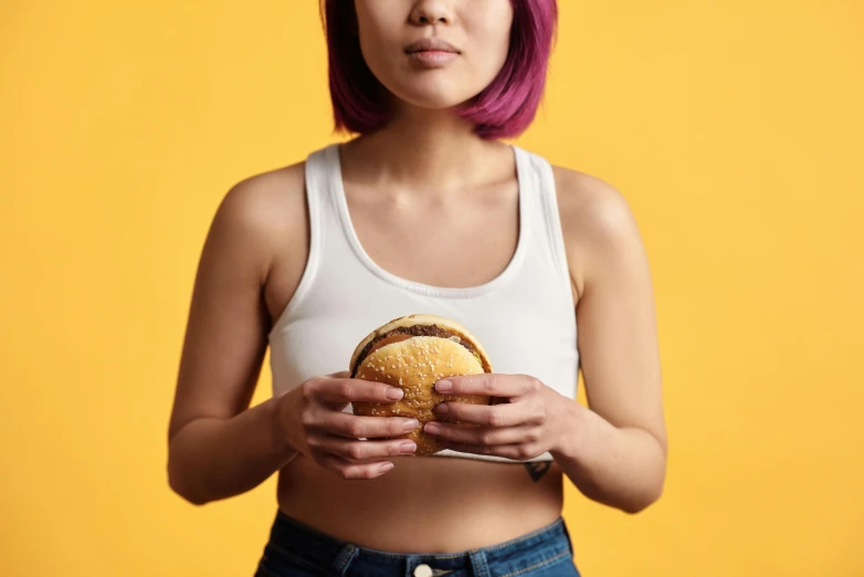 a woman holding a hamburger in front of her face, trending on pexels, hyperrealism, wearing yellow croptop, purple halter top, a young asian woman, wearing a muscle tee shirt