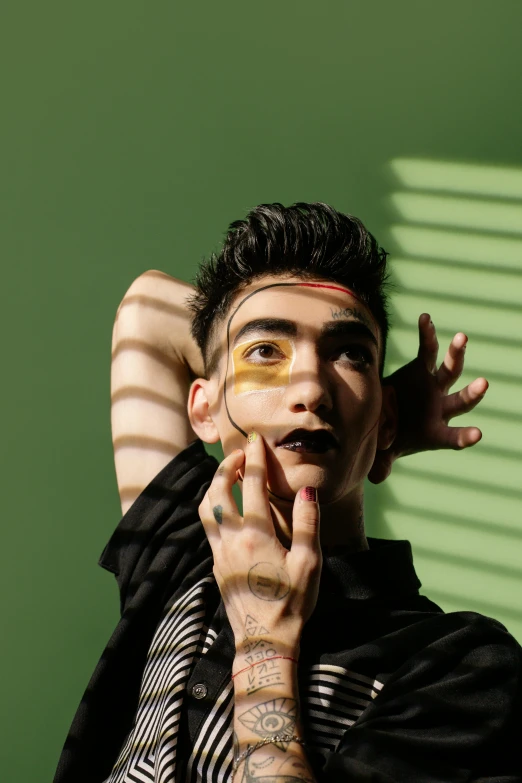 a man standing in front of a green wall, an album cover, inspired by Taro Yamamoto, trending on pexels, holding his hands up to his face, attractive androgynous humanoid, stripe over eye, diego fernandez