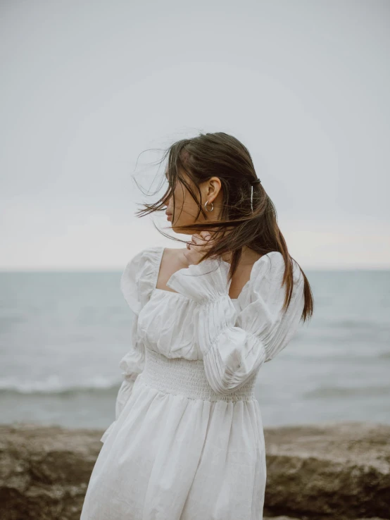 a woman standing on top of a beach next to the ocean, inspired by Wilhelm Hammershøi, pexels contest winner, romanticism, her hair is white, long sleeves, young asian girl, puff sleeves