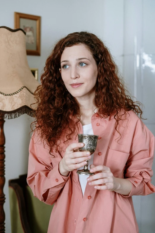 a woman holding a wine glass in front of a lamp, inspired by Anita Malfatti, pexels contest winner, renaissance, curly copper colored hair, wearing a light - pink suit, holding a bottle of arak, is ((drinking a cup of tea))