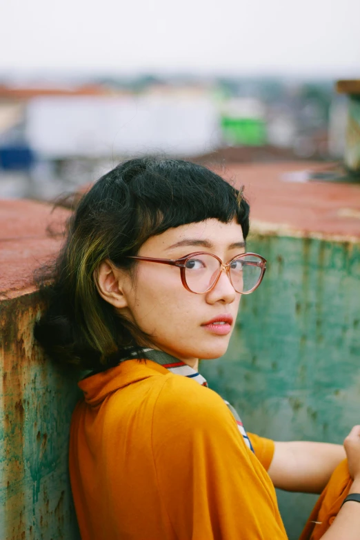 a woman wearing glasses leaning against a wall, a picture, inspired by Ruth Jên, trending on pexels, realism, on a boat, square glasses, vietnamese woman, curly bangs