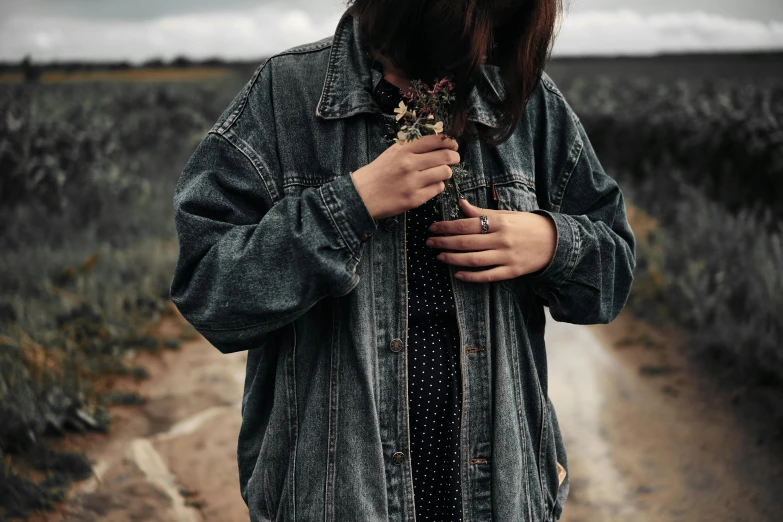 a woman standing in the middle of a dirt road, an album cover, trending on pexels, realism, jean jacket, overgrown with flowers, gray skin. grunge, detailed clothing