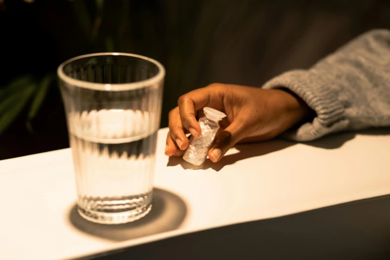 a person holding a piece of ice next to a glass of water, pills, low lighting, lena oxton, on a table