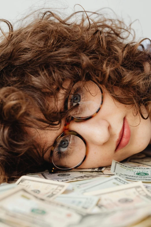 a woman laying on top of a pile of money, trending on pexels, renaissance, brown curly hair, square rimmed glasses, teenage boy, bedhead