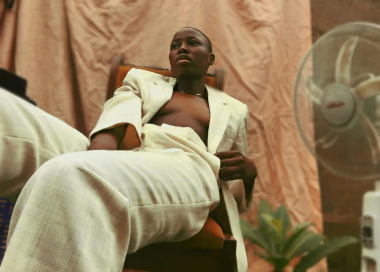 a man sitting in a chair in front of a fan, an album cover, trending on pexels, lupita nyong'o, tan suit, masculine appeal high fashion, androgynous