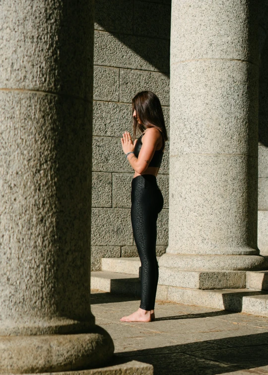 a woman standing in front of a stone building, by Nina Hamnett, pexels contest winner, arabesque, marjaryasana and bitilasana, black spandex, praying posture, profile image