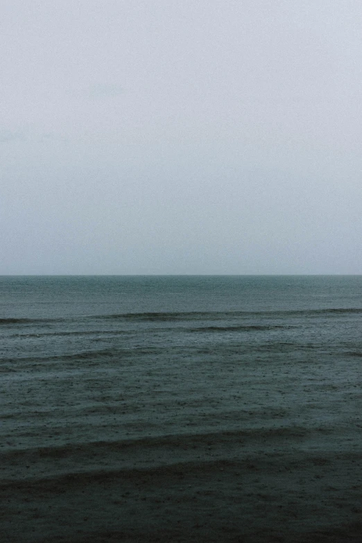 a man standing on top of a beach next to the ocean, inspired by Andreas Gursky, postminimalism, overcast gray skies, 35mm —w 1920 —h 1080, muted green, landscape photograph