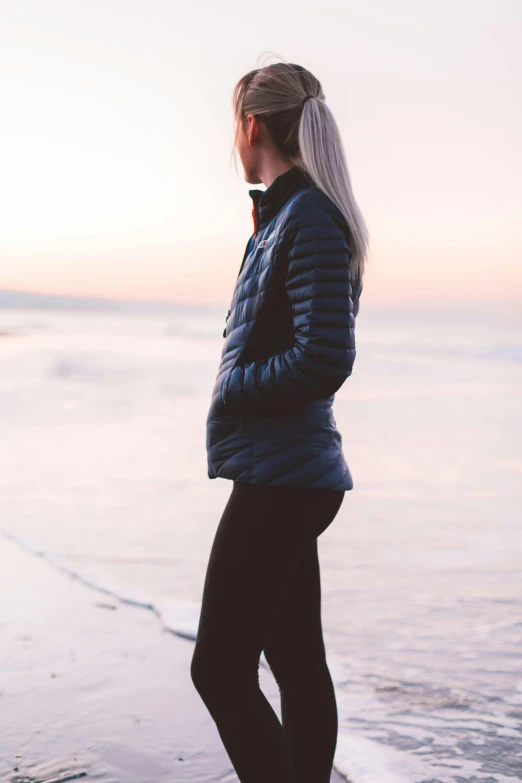 a woman standing on top of a beach next to the ocean, inspired by Louisa Matthíasdóttir, unsplash, model wears a puffer jacket, full body profile, wearing fitness gear, beatifully lit