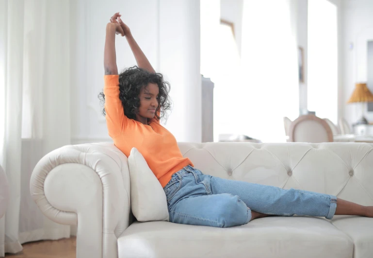 a woman sitting on top of a white couch, pexels contest winner, happening, wearing an orange t shirt, arms stretched out, wearing jeans, young black woman