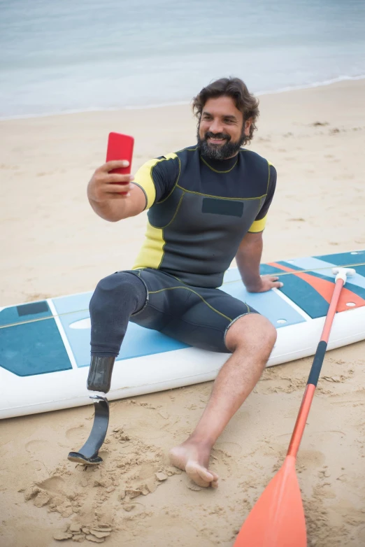 a man sitting on top of a surfboard on a beach, wearing bionic implants, hold up smartphone, square, crutches