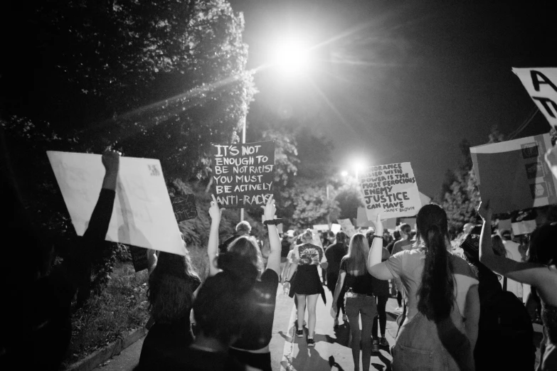 a group of people walking down a street holding signs, a black and white photo, by Emma Andijewska, pexels, summer night, protest, against the backdrop of trees, jordan lamarre - wan