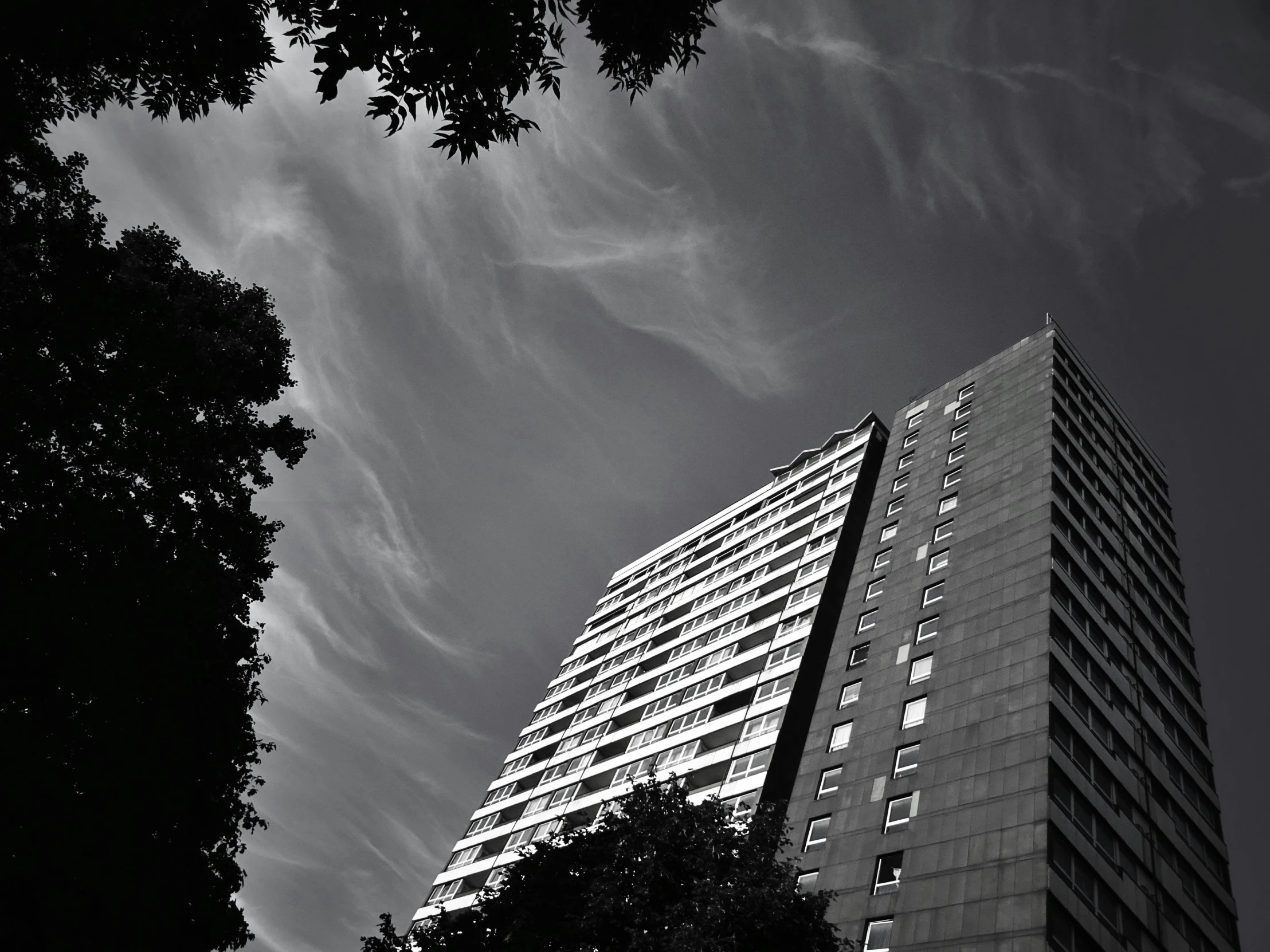 a black and white photo of a tall building, a black and white photo, inspired by Richard Wilson, summer sky, looming trees, concrete housing, dark towering clouds