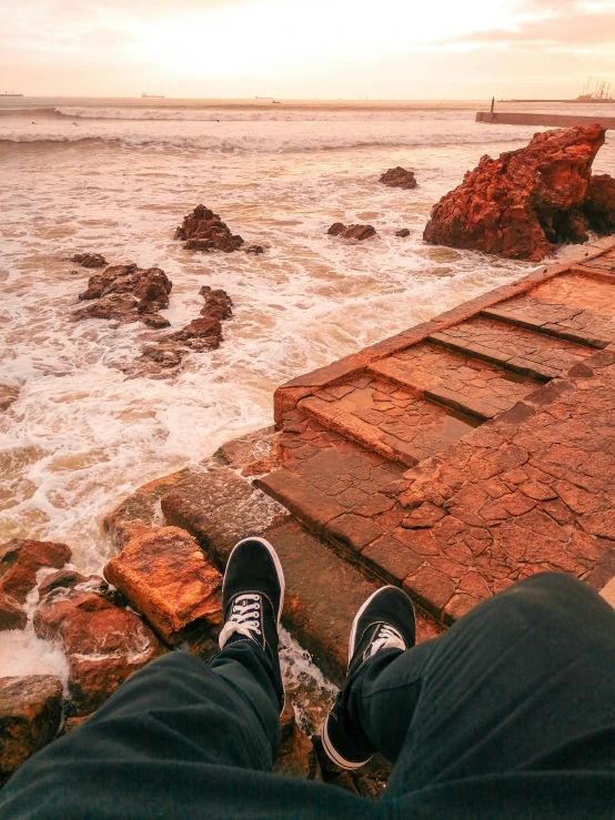 a person sitting on the edge of a pier next to the ocean, pexels contest winner, realism, red sand, waves crashing at rocks, sneaker photo, sitting on temple stairs