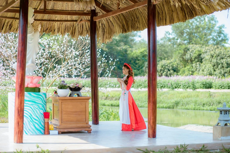 a woman in a red and white dress standing under a gazebo, inspired by Cui Bai, ao dai, tea ceremony scene, thatched roof, spa