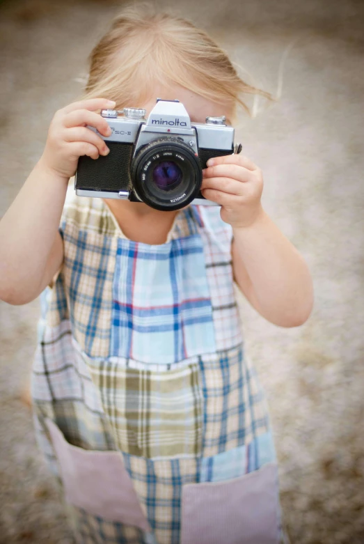 a little girl taking a picture with a camera, inspired by Vivian Maier, unsplash, blue checkerboard dress, multicoloured, grey, small