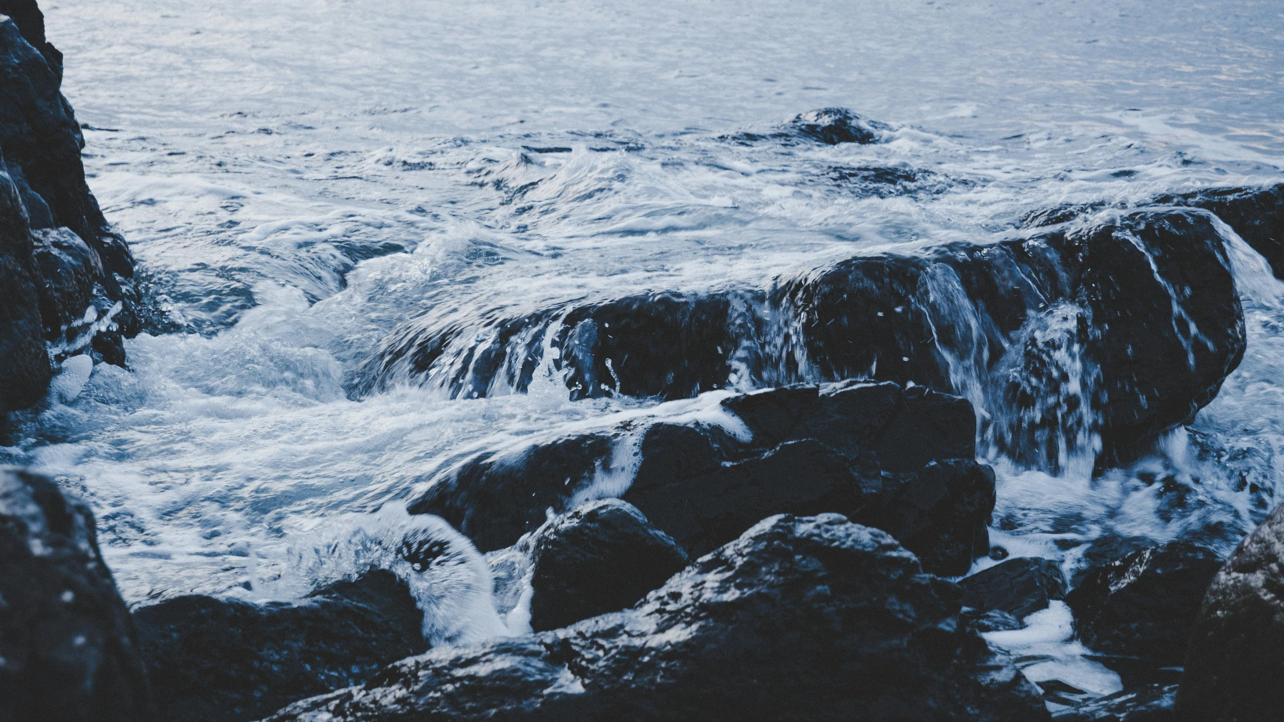 a man standing on top of a rock next to the ocean, an album cover, by Christen Dalsgaard, unsplash, rushing water, blue toned, with lots of dark grey rocks, thumbnail