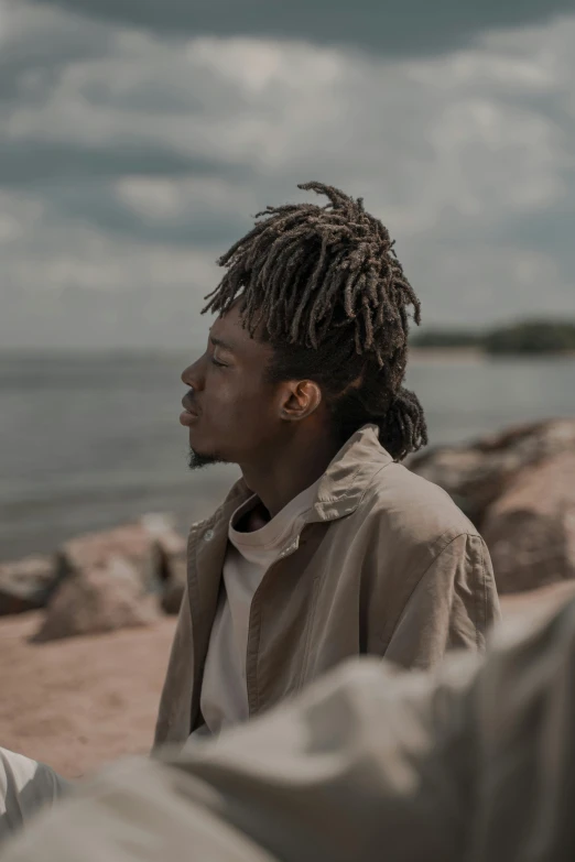 a man sitting on a beach next to a body of water, an album cover, pexels contest winner, dreadlock breed hair, headshot profile picture, ( ( theatrical ) ), muted browns