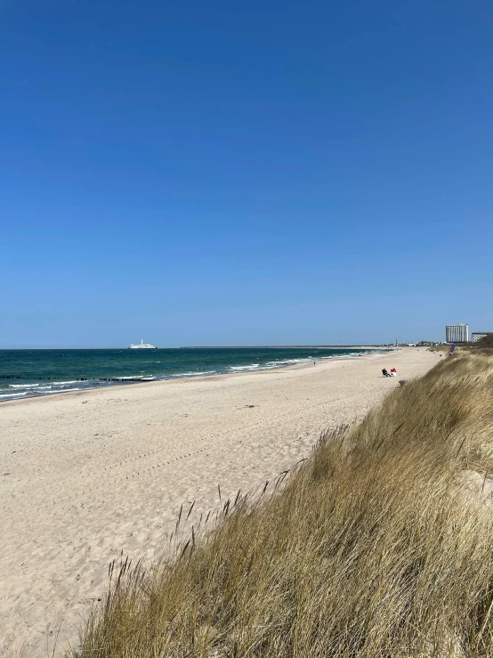 a sandy beach next to the ocean on a sunny day, by Niko Henrichon, happening, building in the distance, hannover, clear skies in the distance, a green