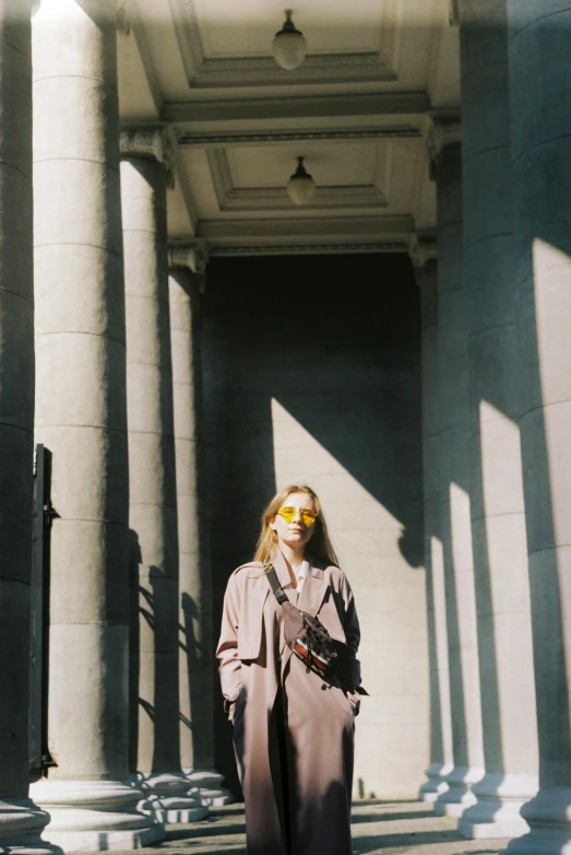 a woman standing in front of a building, harsh sun light, vhs colour photography, marble columns in background, woman is in a trenchcoat