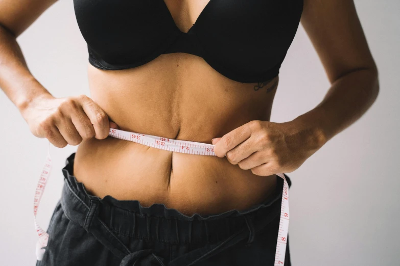 a woman measuring her waist with a tape, by Nicolette Macnamara, trending on pexels, manuka, belly button showing, looking towards camera, lean and toned