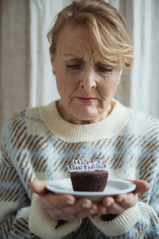 a woman holding a plate with a cupcake on it, aging, quiet intensity, he is wearing a brown sweater, birthday cake