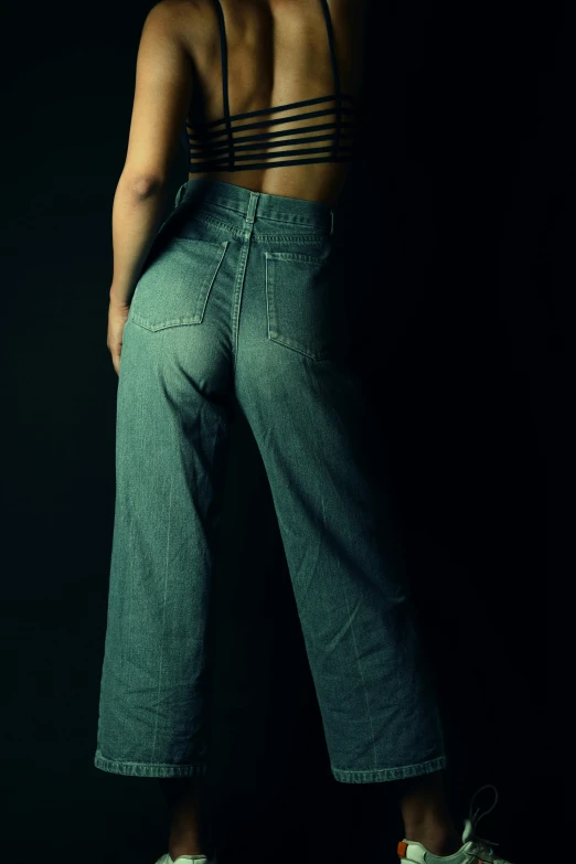 a woman standing with her back to the camera, an album cover, inspired by Elsa Bleda, trending on pexels, hyperrealism, jean pants, with a black background, dirty green clothes, large pants