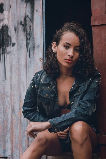 a woman sitting on top of a wooden bench, by Andrée Ruellan, pexels contest winner, photorealism, curly middle part haircut, alone gorgeous latin woman, jean jacket, leaning on door