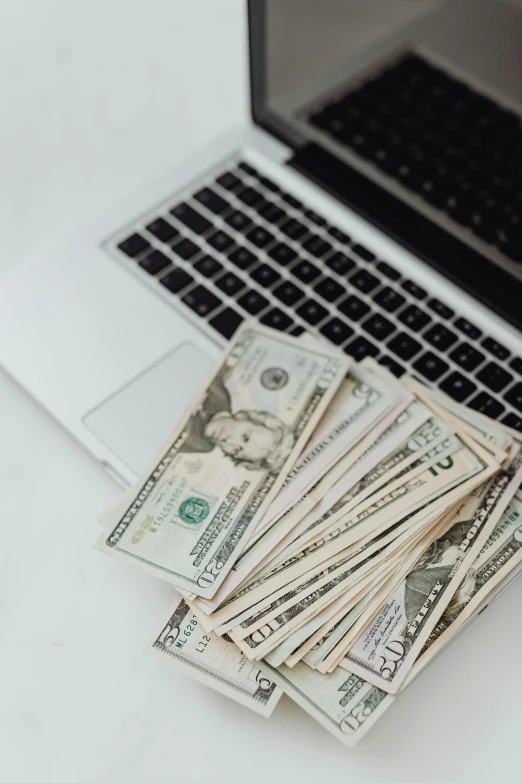 a laptop computer sitting next to a pile of money, by Byron Galvez, trending on unsplash, 10k, plain background, multiple stories, 8