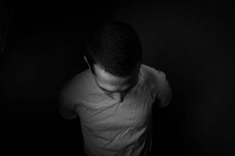 a man in a white shirt standing in a dark room, a black and white photo, by Adam Marczyński, pexels, top-down shot, he is sad, avatar image, top down shot