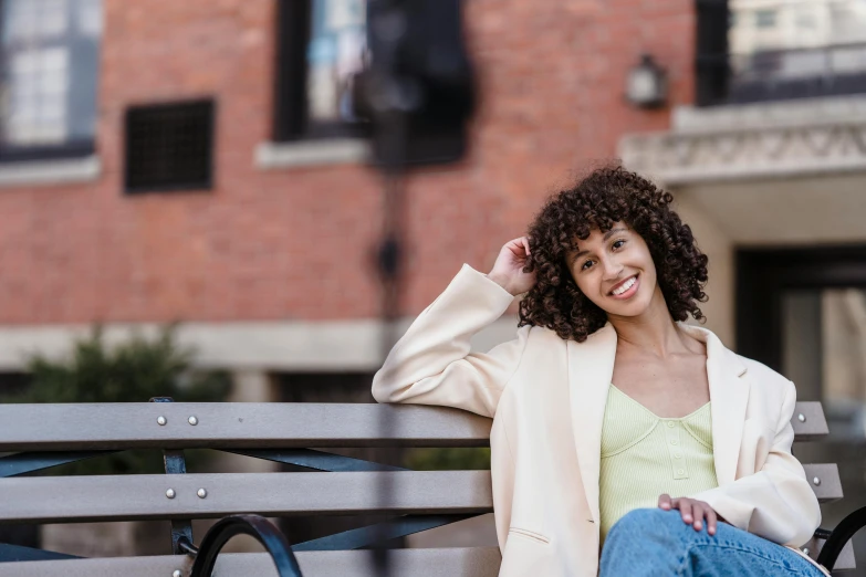 a woman sitting on a bench in front of a building, by Washington Allston, trending on pexels, brown curly hair, promotional image, wesley kimler, posing for the camera