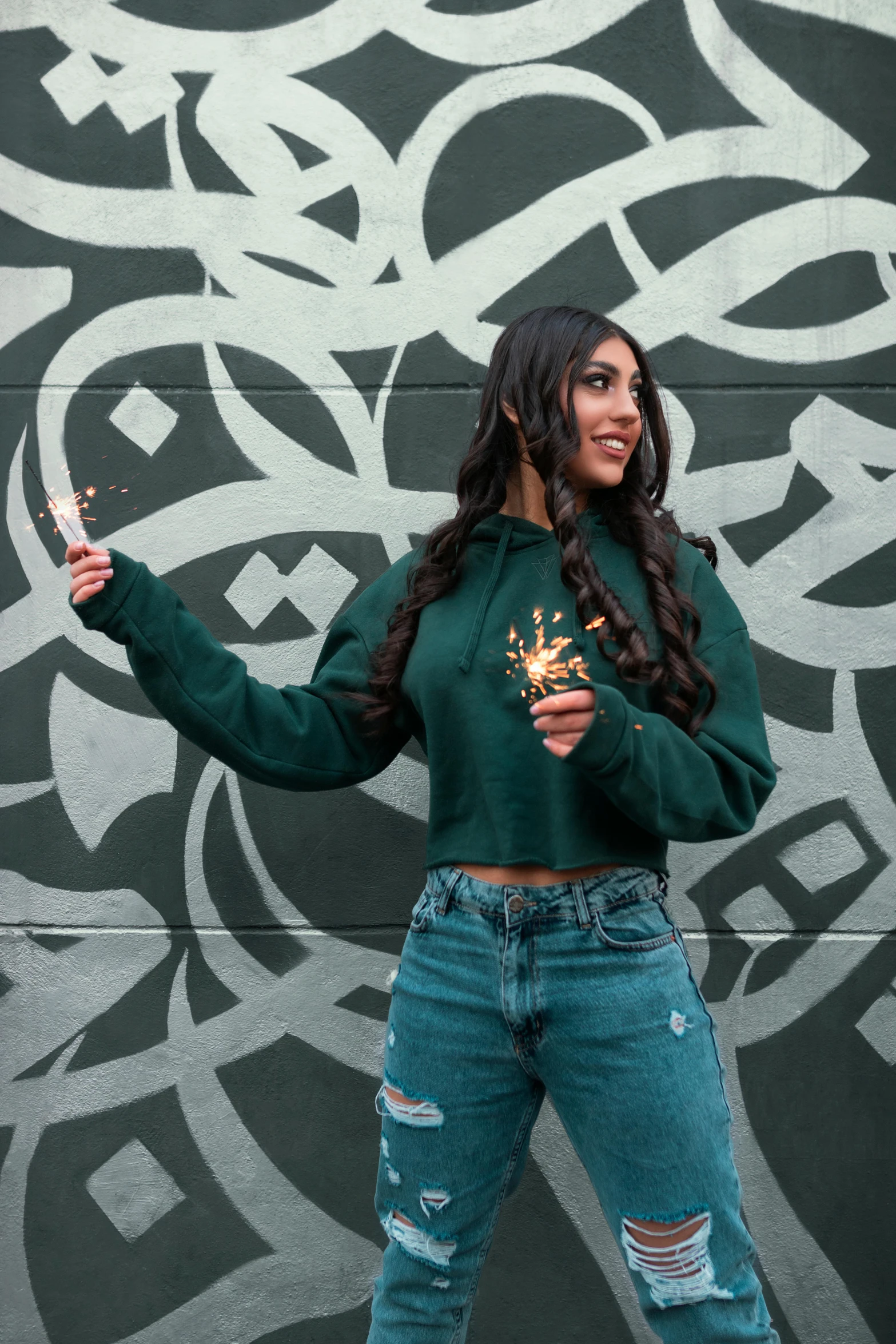 a woman standing in front of a graffiti wall, inspired by Pia Fries, trending on pexels, ornate dark green clothing, arab, wearing sweatshirt, holding a bottle