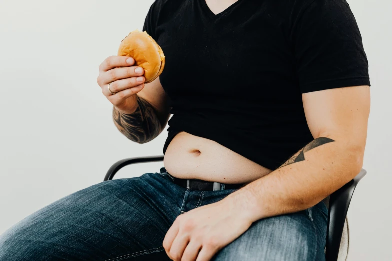 a man sitting on a chair eating a donut, trending on pexels, her belly is fat and round, jeans and t shirt, non-binary, swollen muscles