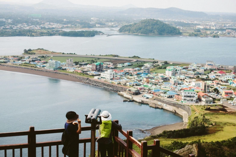 a couple of people standing on top of a hill, a tilt shift photo, mingei, the sea seen behind the city, travel guide, te pae, panoramic view of girl