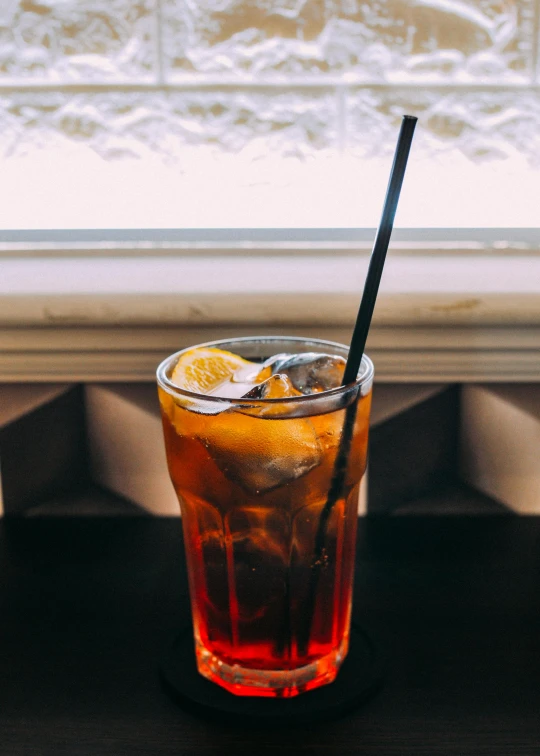 a drink sitting on top of a table next to a window, a portrait, by Matt Cavotta, unsplash, tall iced tea glass, black and orange, with a straw, half - length photo