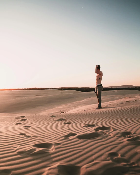 a man standing on top of a sandy beach, inspired by Russell Drysdale, unsplash contest winner, desert mirage, slightly tanned, sun down, waving