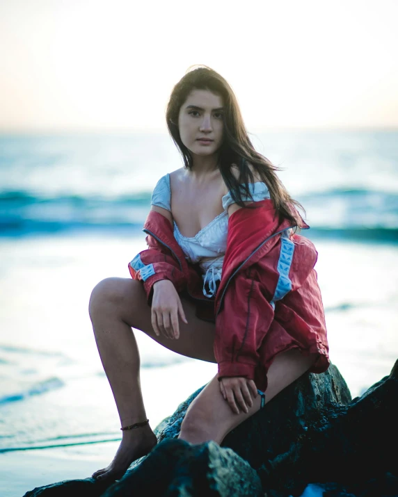 a woman sitting on a rock on the beach, an album cover, inspired by Elsa Bleda, unsplash, renaissance, wearing red jacket, mia khalifa, wearing a white bikini, 8k octan photo