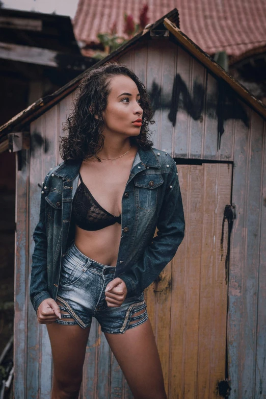 a woman standing in front of a wooden shed, by Andrée Ruellan, pexels contest winner, sexy girl wearing shorts, wearing a jeans jackets, mixed-race woman, in a black betch bra