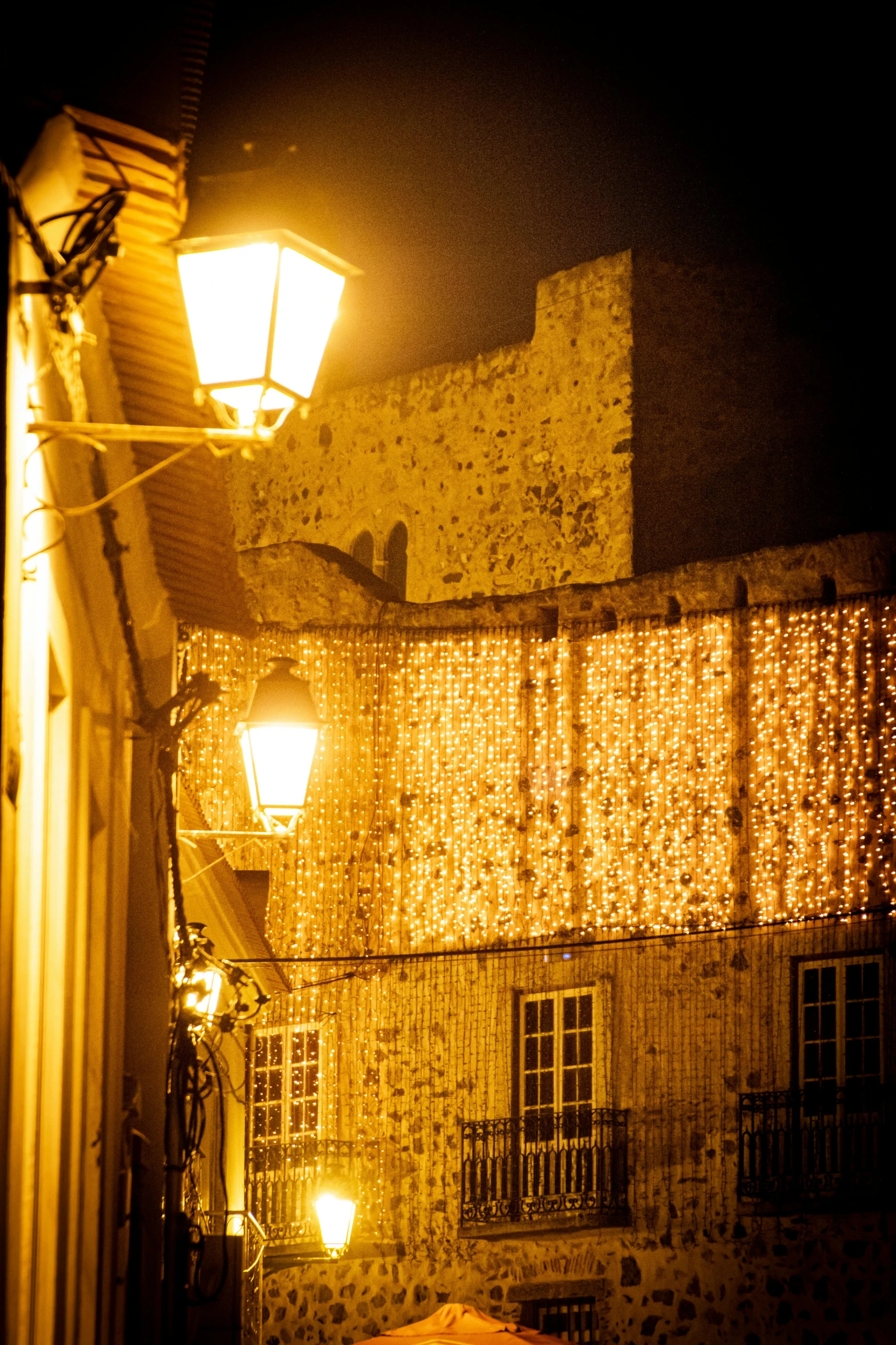 a couple of people walking down a street at night, renaissance, gold decorations, city wall, nazare (portugal), string lights