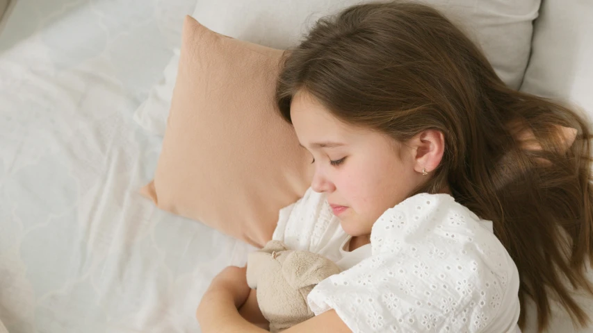 a little girl laying in bed with a teddy bear, inspired by Sarah Lucas, light beige pillows, close up shot from the side, easy to use, bedhead