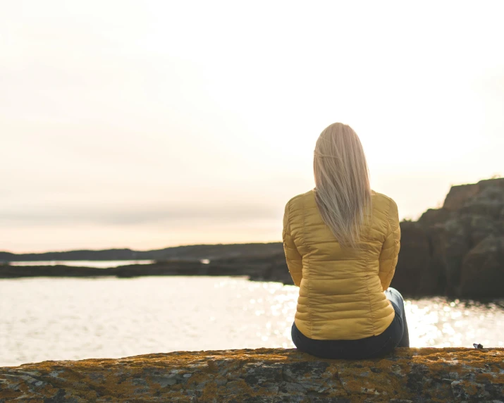 a woman sitting on a rock overlooking a body of water, inspired by Louisa Matthíasdóttir, unsplash, yellow hue, looking onto the horizon, back - lit, blonde swedish woman