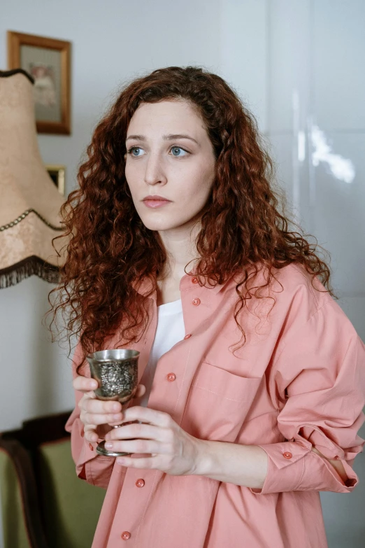 a woman in a pink shirt holding a wine glass, by Isabel Naftel, pexels, renaissance, curly copper colored hair, holding a candle holder, looking serious, wearing pajamas